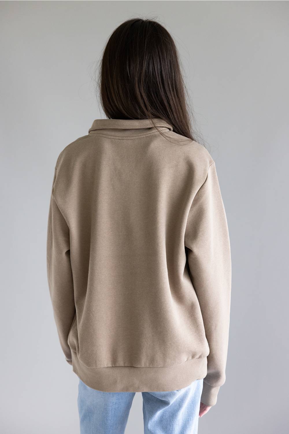 A person viewed from behind wearing a light beige crewneck sweatshirt and light blue jeans against a plain gray studio background.