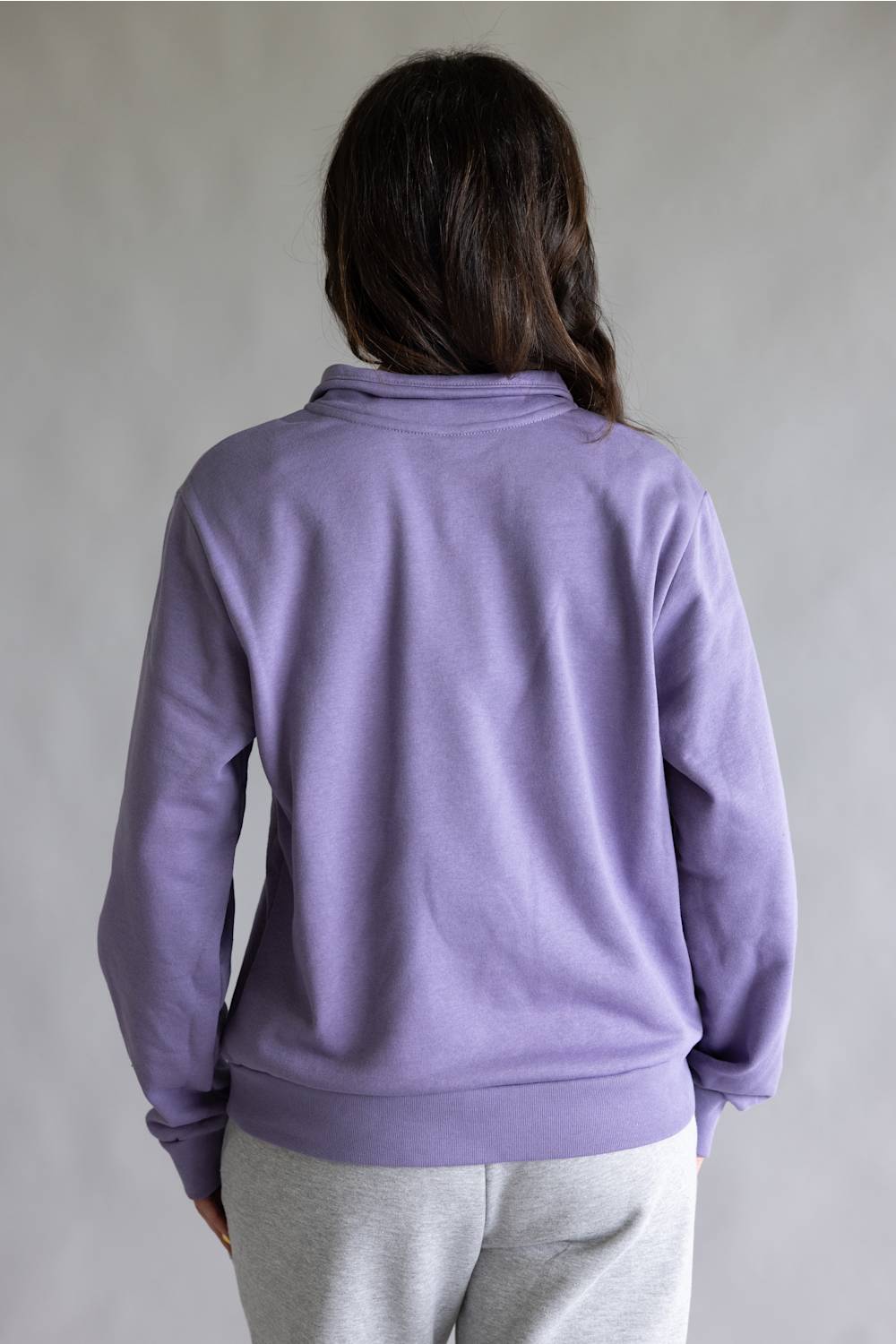 A person shown from the back wearing a lavender-purple long-sleeve sweatshirt and light gray sweatpants against a plain gray studio background.