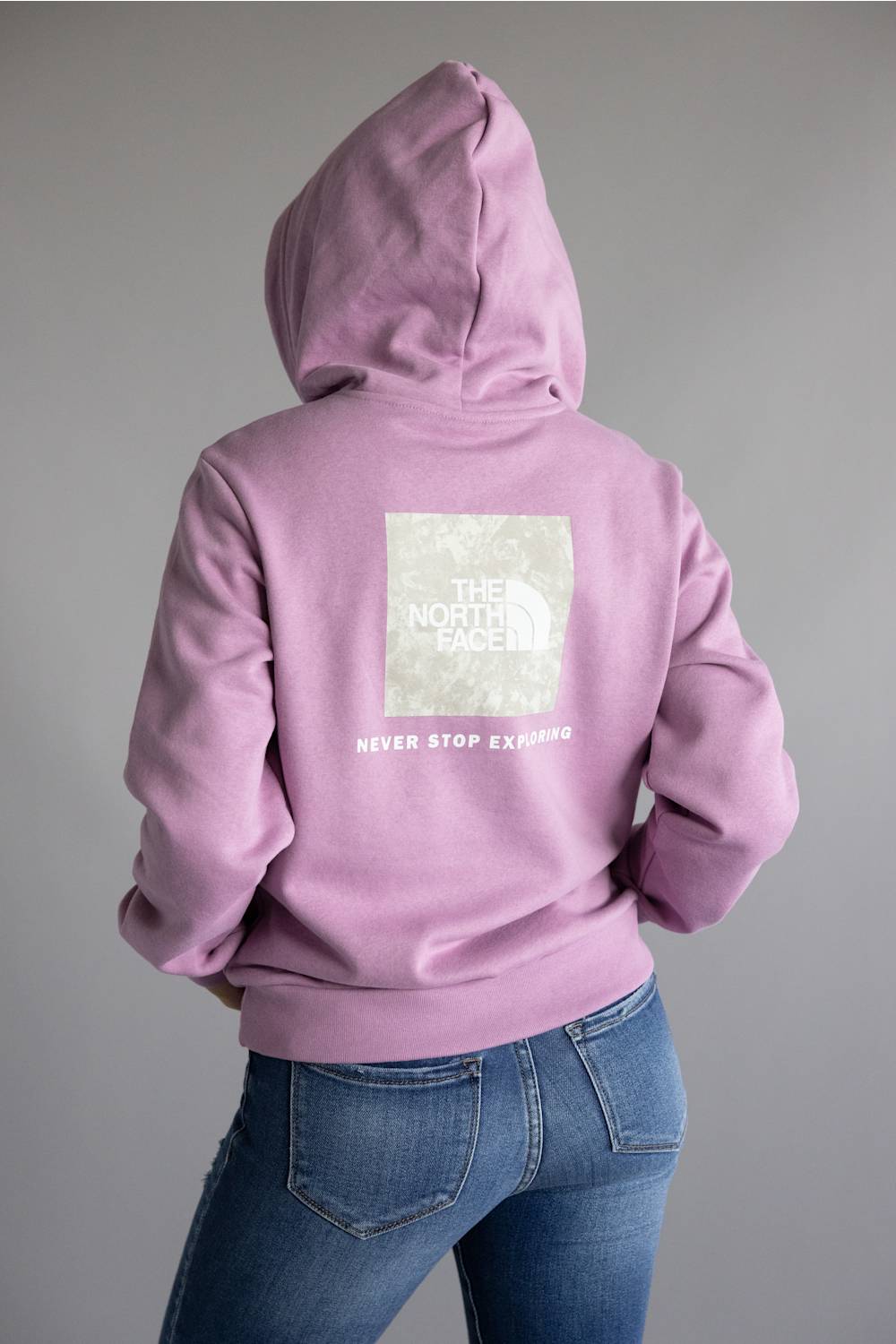 A person shown from the back wearing a lavender-purple hoodie with a large white The North Face logo patch on the back, paired with blue jeans, against a plain gray studio background.