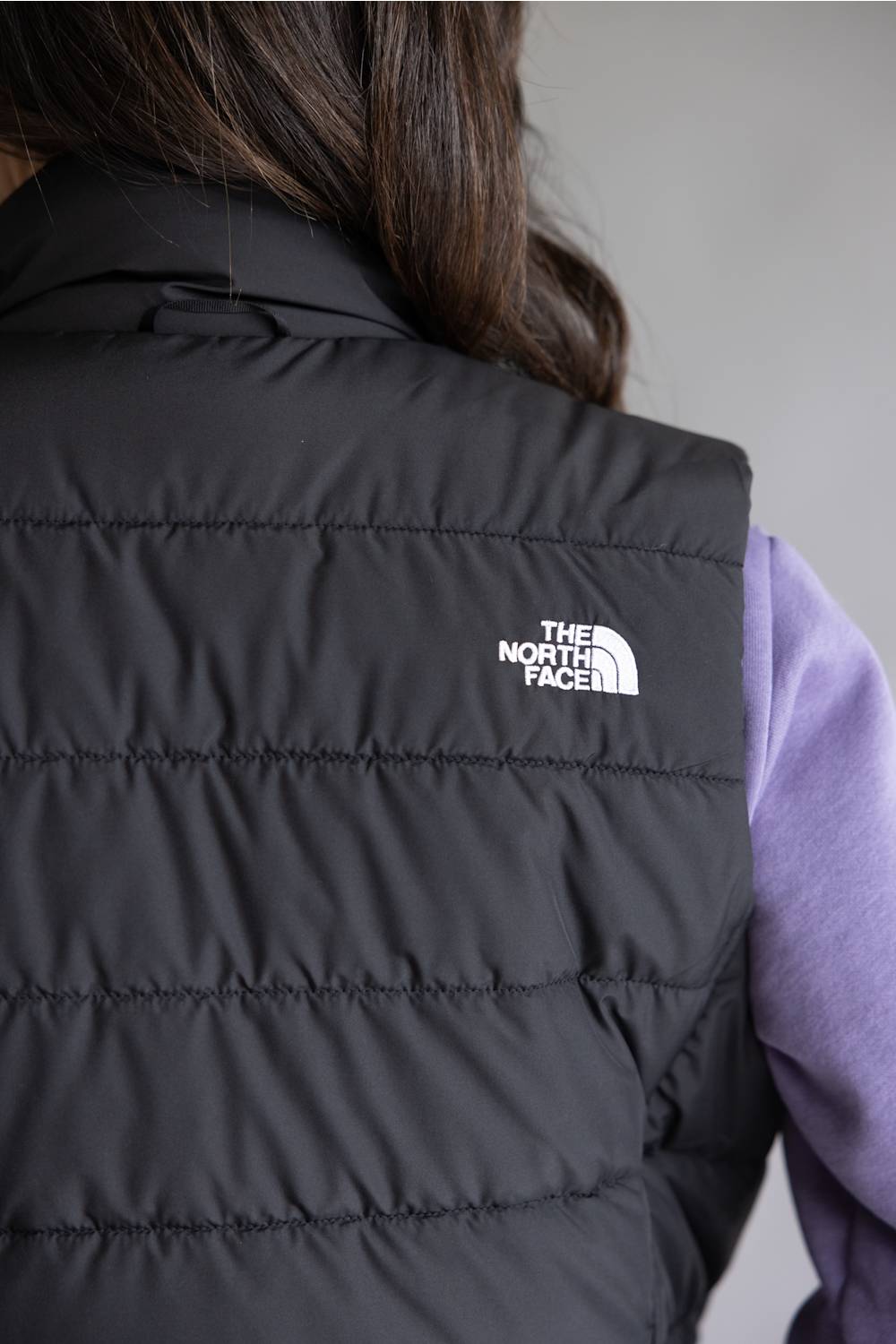 Close-up back view of a person wearing a black quilted puffer vest with a white The North Face logo on the upper back, layered over a light purple long-sleeve shirt.