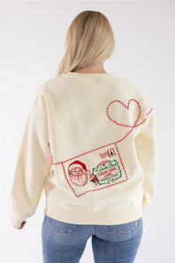 A woman viewed from the back wearing a cream-colored sweatshirt with a large red and green Christmas-themed envelope graphic on the back, paired with blue jeans, against a plain white background.