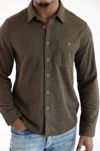 A man wearing a dark brown textured long-sleeve button-up shirt with a chest pocket and light blue jeans, shown from the neck down against a white background.