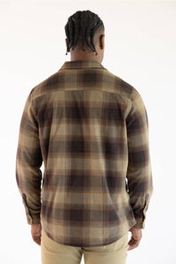A man viewed from behind wearing a long-sleeve plaid flannel shirt in brown, beige, and black, paired with khaki pants, against a plain white background.