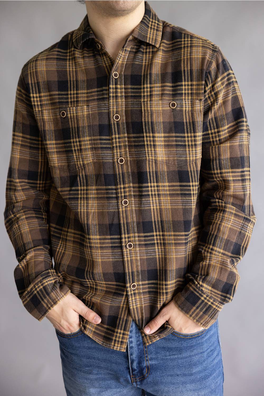 A man wearing a brown and black plaid button-down shirt with blue jeans, standing against a neutral gray background.