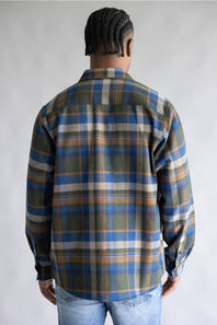 A man viewed from behind wearing a long-sleeve plaid button-up shirt in blue, olive green, tan, and white, paired with light wash blue jeans against a neutral gray studio background.