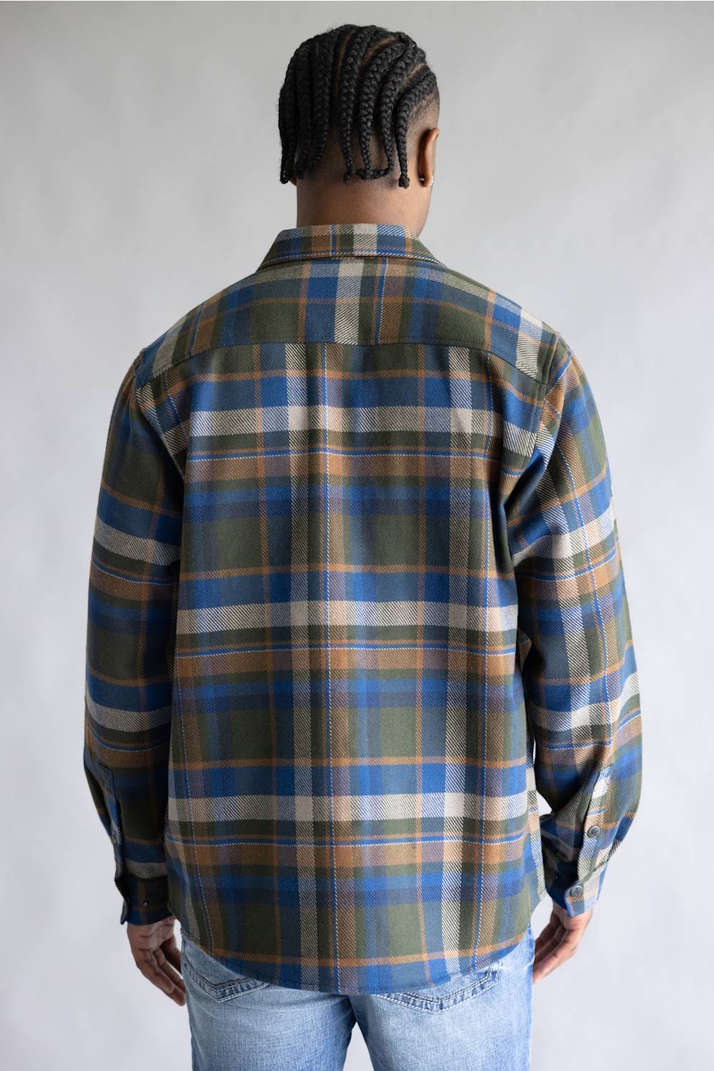 A man viewed from behind wearing a long-sleeve plaid button-up shirt in blue, olive green, tan, and white, paired with light wash blue jeans against a neutral gray studio background.