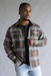 A man wearing a brown and beige plaid flannel shirt jacket over a black henley t-shirt and light wash jeans, standing against a neutral gray studio background.