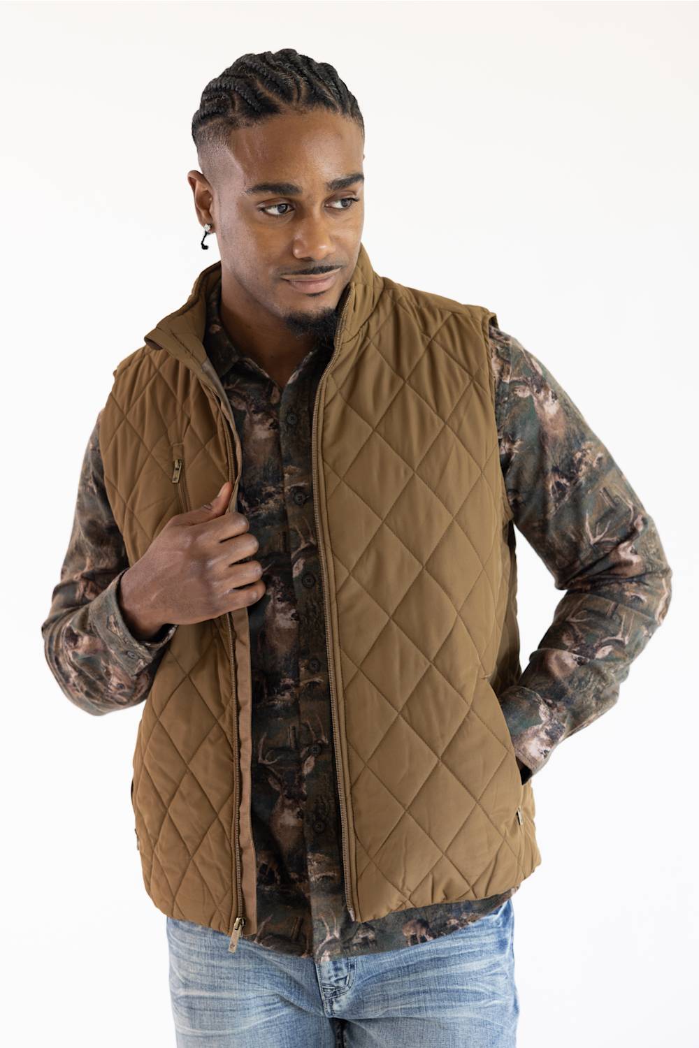 A man wearing a brown diamond-quilted zip-up vest over a camouflage long-sleeve shirt and blue jeans, photographed against a plain white studio background.