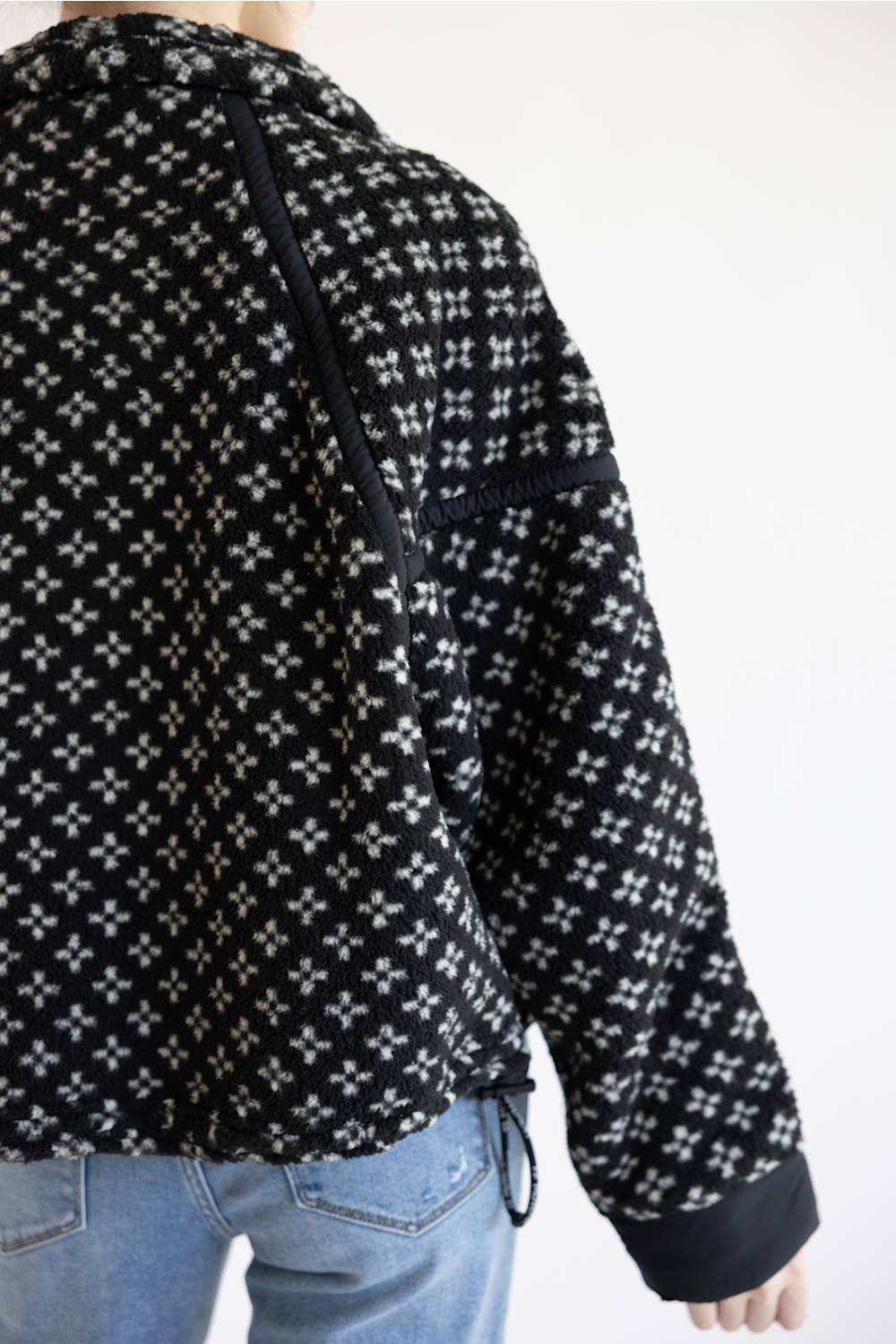 A person wearing a black pullover with a white geometric cross pattern, paired with light blue jeans, shown from the back against a plain white background.