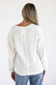 A woman viewed from the back wearing a white ribbed knit sweater with a decorative bow detail at the neckline and blue jeans, against a plain white background.