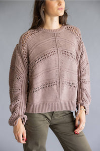 A woman wearing a dusty mauve knit sweater with textured openwork patterns and olive green pants, shown from the head down against a plain gray studio background.