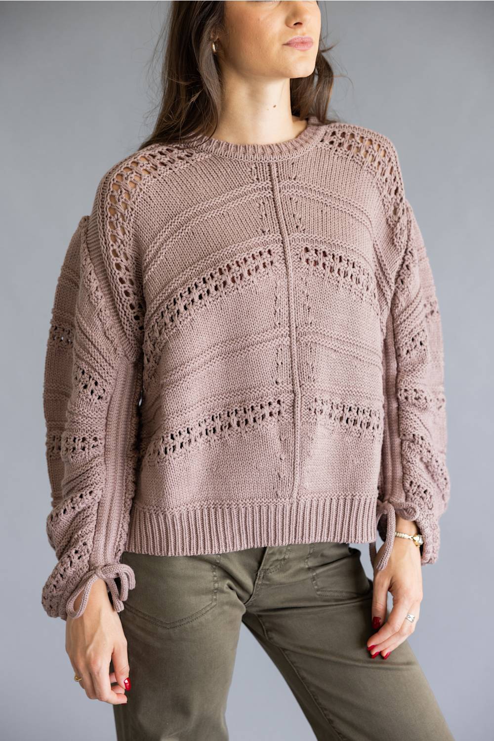A woman wearing a dusty mauve knit sweater with textured openwork patterns and olive green pants, shown from the head down against a plain gray studio background.