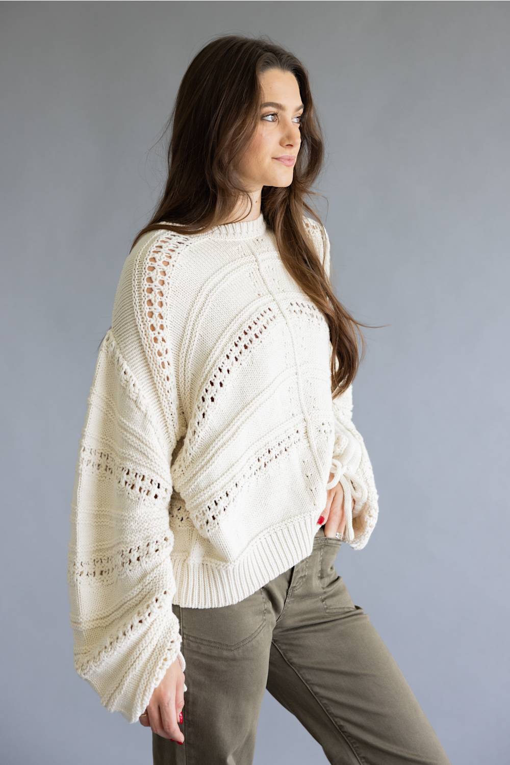 A woman wearing a cream-colored, textured knit sweater with intricate openwork patterns and long sleeves, paired with olive green pants, posed against a solid gray studio background.