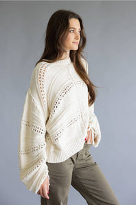 A woman wearing a cream-colored, textured knit sweater with intricate openwork patterns and long sleeves, paired with olive green pants, posed against a solid gray studio background.
