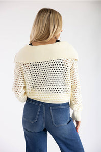 A woman shown from the back wearing a cream-colored open-knit cropped sweater with a wide ribbed collar and blue jeans, against a plain white studio background.