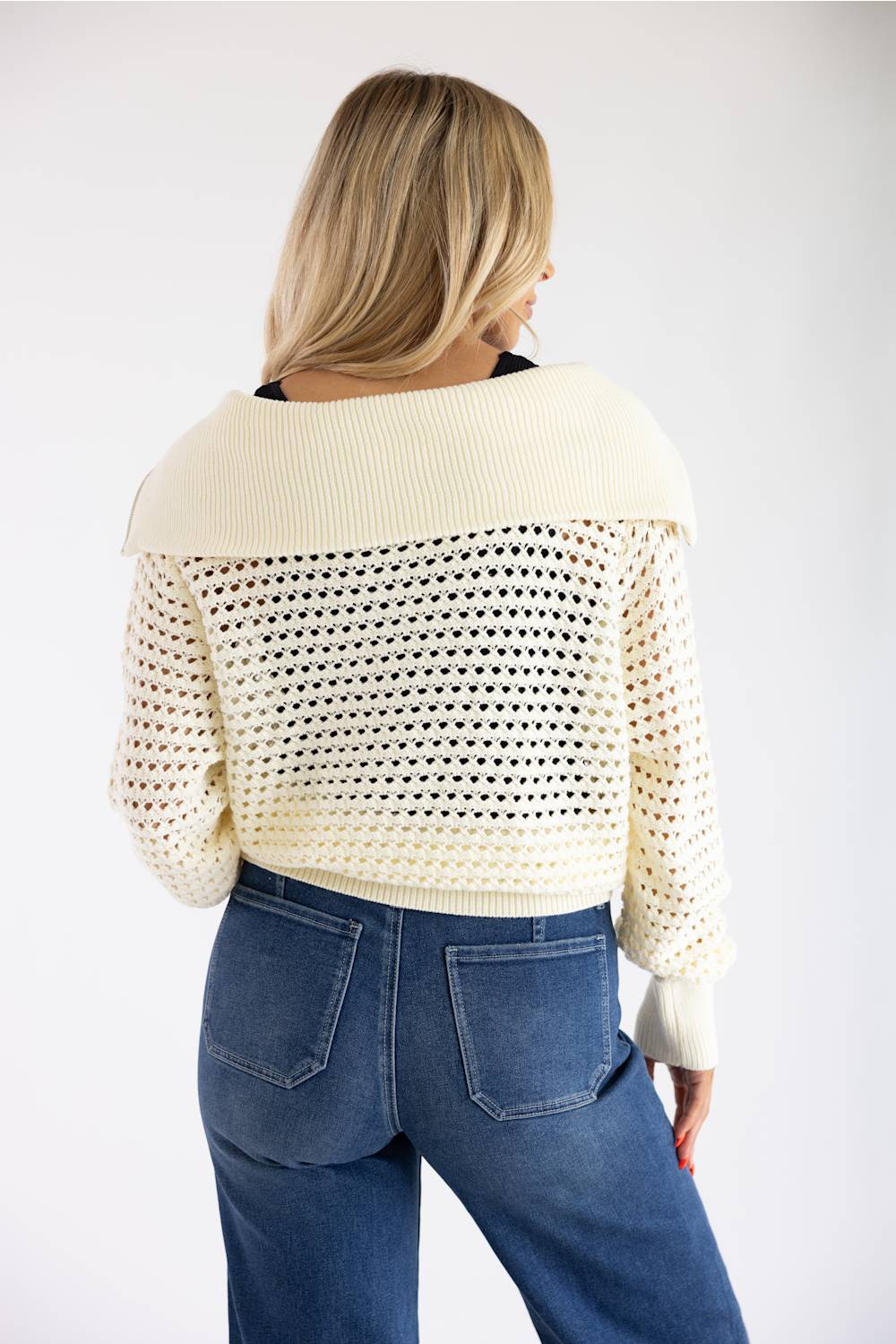 A woman shown from the back wearing a cream-colored open-knit cropped sweater with a wide ribbed collar and blue jeans, against a plain white studio background.