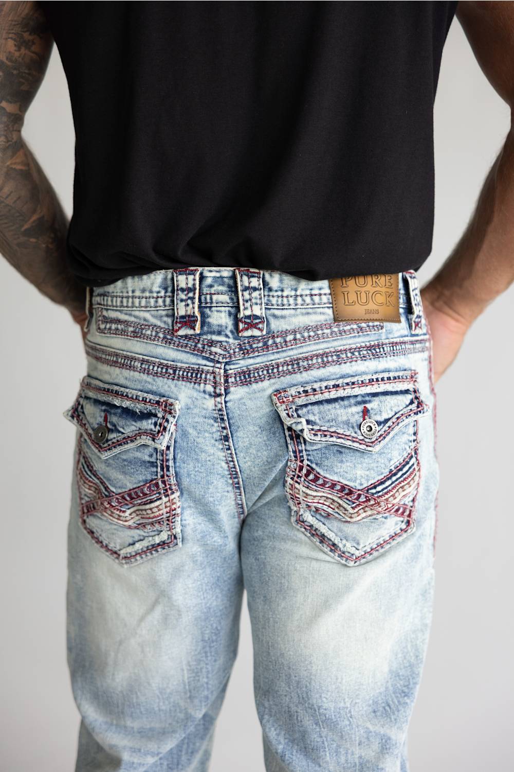 Rear view of a person wearing light-wash blue denim jeans with heavily contrasted red and black embroidery on the back pockets, paired with a black t-shirt against a light gray background.