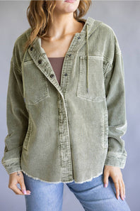 A woman wearing an oversized corduroy button-up shirt in sage green, styled over a brown top and blue jeans against a neutral gray studio background.