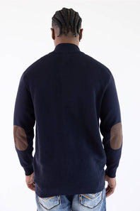 Man shown from the back wearing a navy mock neck jacket with brown elbow patches standing against a white background