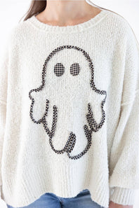 A person wearing a chunky off-white knit sweater with a black-and-light orange gingham ghost applique on the front, paired with blue jeans against a plain white background.