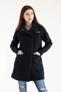 A woman with red hair wearing a black fleece coat and blue jeans, standing against a plain white studio background. The coat has a small white embroidered 'Columbia' logo on the left chest.