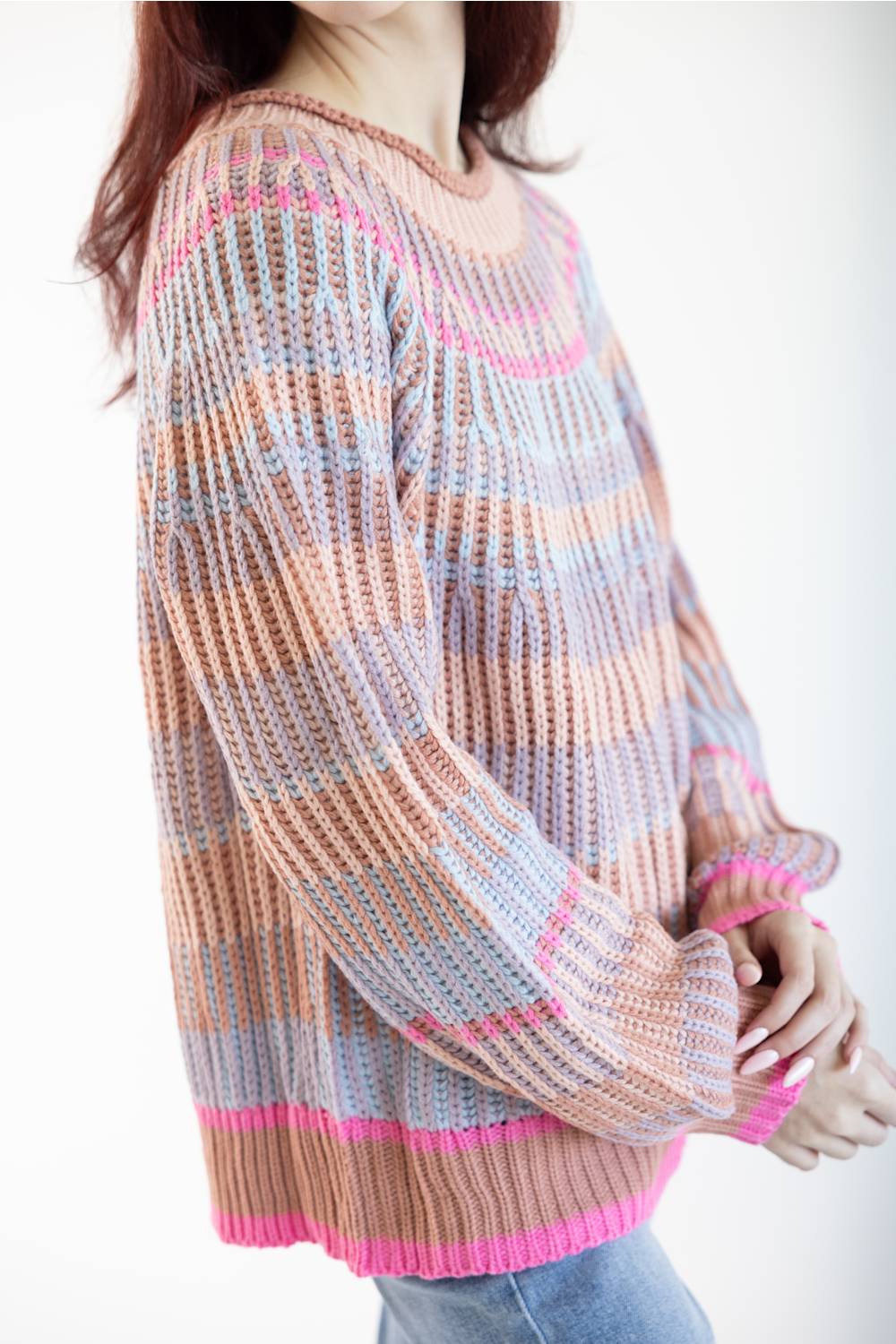 A woman wearing a chunky knit sweater with horizontal stripes in peach, light blue, and bright pink, paired with light blue jeans against a plain white background.