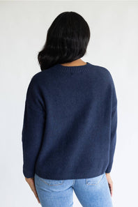A woman viewed from behind wearing a navy blue knit sweater and light blue jeans against a plain white background.