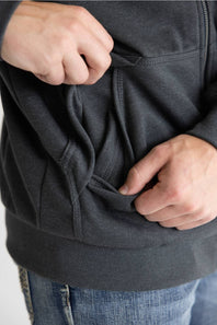 Close-up of a person wearing a dark charcoal gray zip-up hoodie and blue jeans, demonstrating the spacious side pockets by pulling the fabric outward with both hands.
