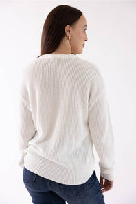 A woman shown from the back wearing a cream-colored ribbed knit sweater and blue jeans against a plain white studio background.