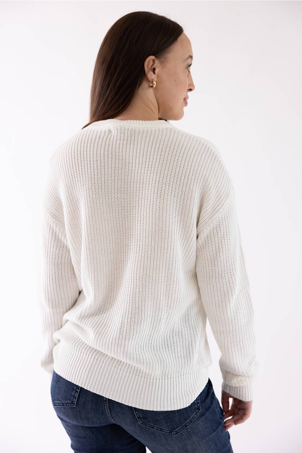 A woman shown from the back wearing a cream-colored ribbed knit sweater and blue jeans against a plain white studio background.
