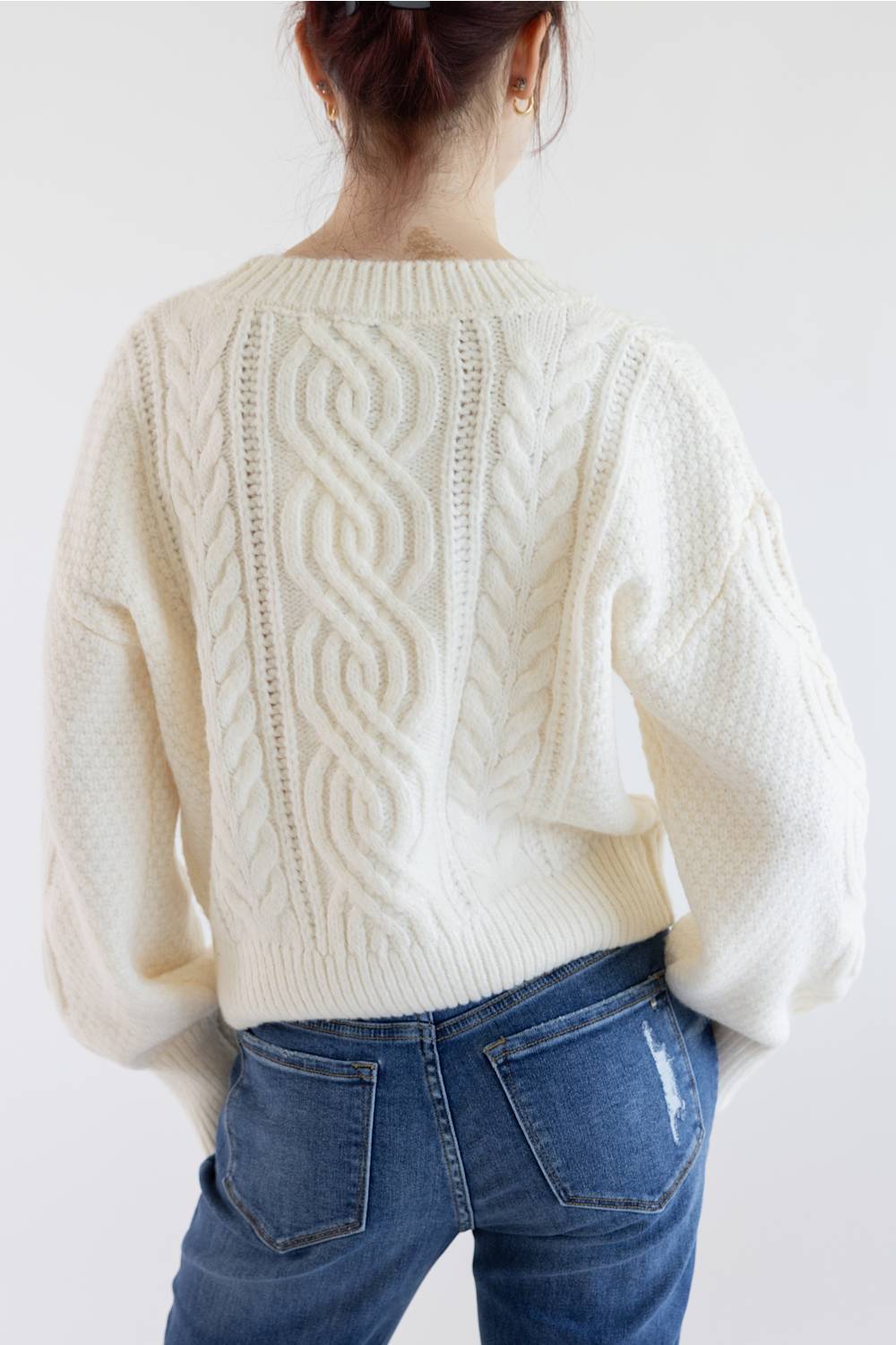 A person shown from the back wearing a cream cable-knit sweater and blue jeans against a plain light background.