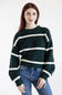 A woman wearing a dark green knit sweater with horizontal cream stripes and blue jeans, standing against a plain white studio background.