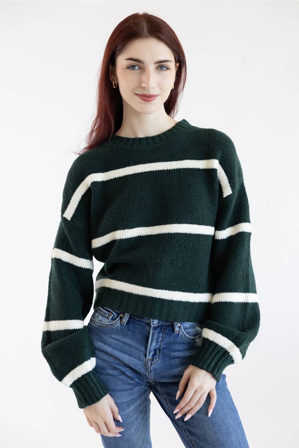 A woman wearing a dark green knit sweater with horizontal cream stripes and blue jeans, standing against a plain white studio background.