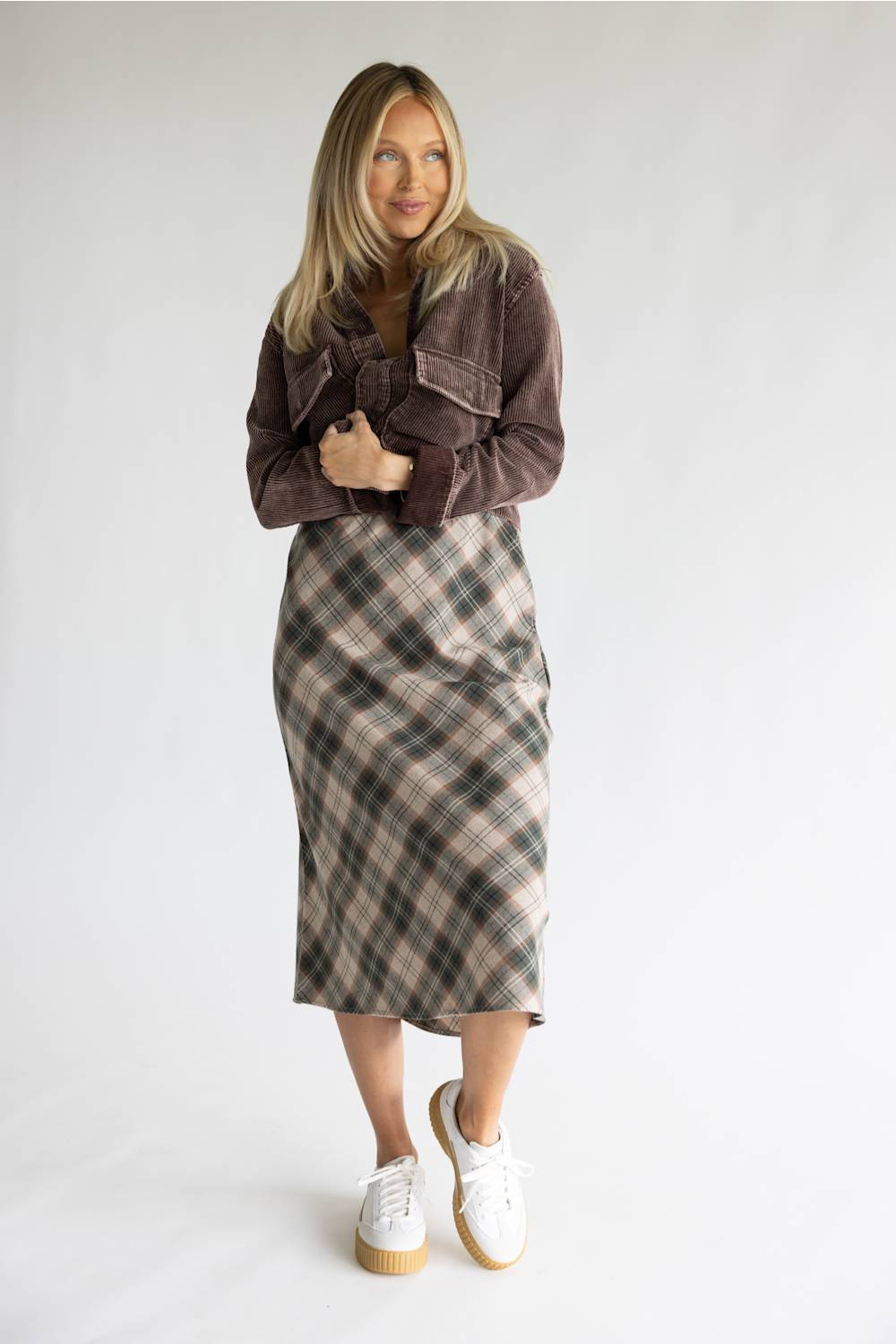 A woman modeling a brown corduroy jacket and a brown, beige, and gray plaid midi skirt, paired with white platform sneakers, against a plain light gray studio background.