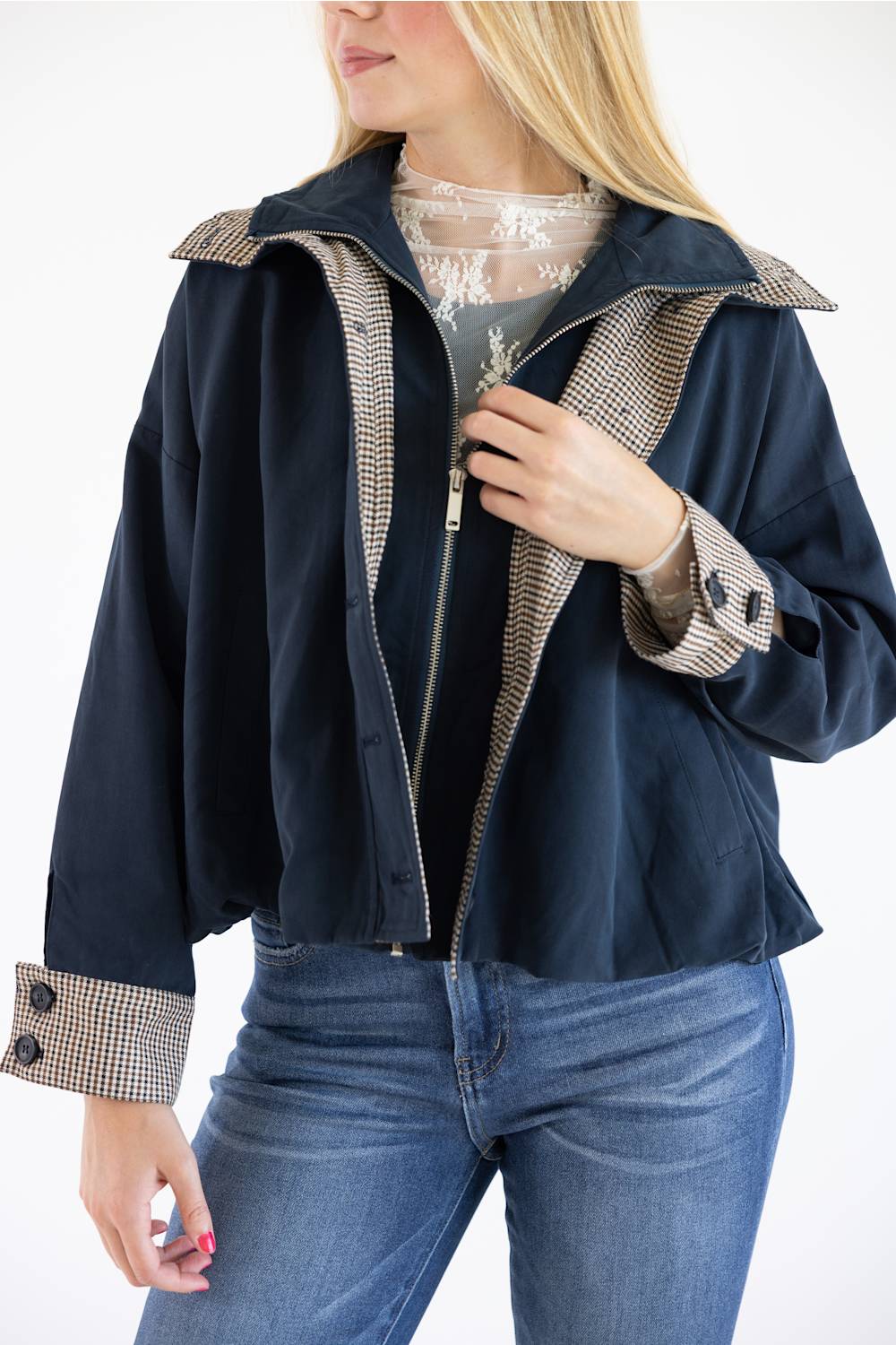 A woman wearing a navy blue jacket with beige and black checkered trim on the collar, cuffs, and front zipper, paired with a lace top and blue jeans.