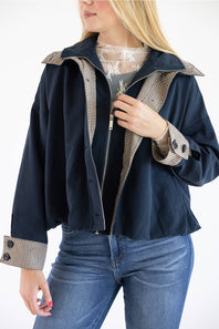 A woman wearing a navy blue jacket with beige and black checkered trim on the collar, cuffs, and front zipper, paired with a lace top and blue jeans.