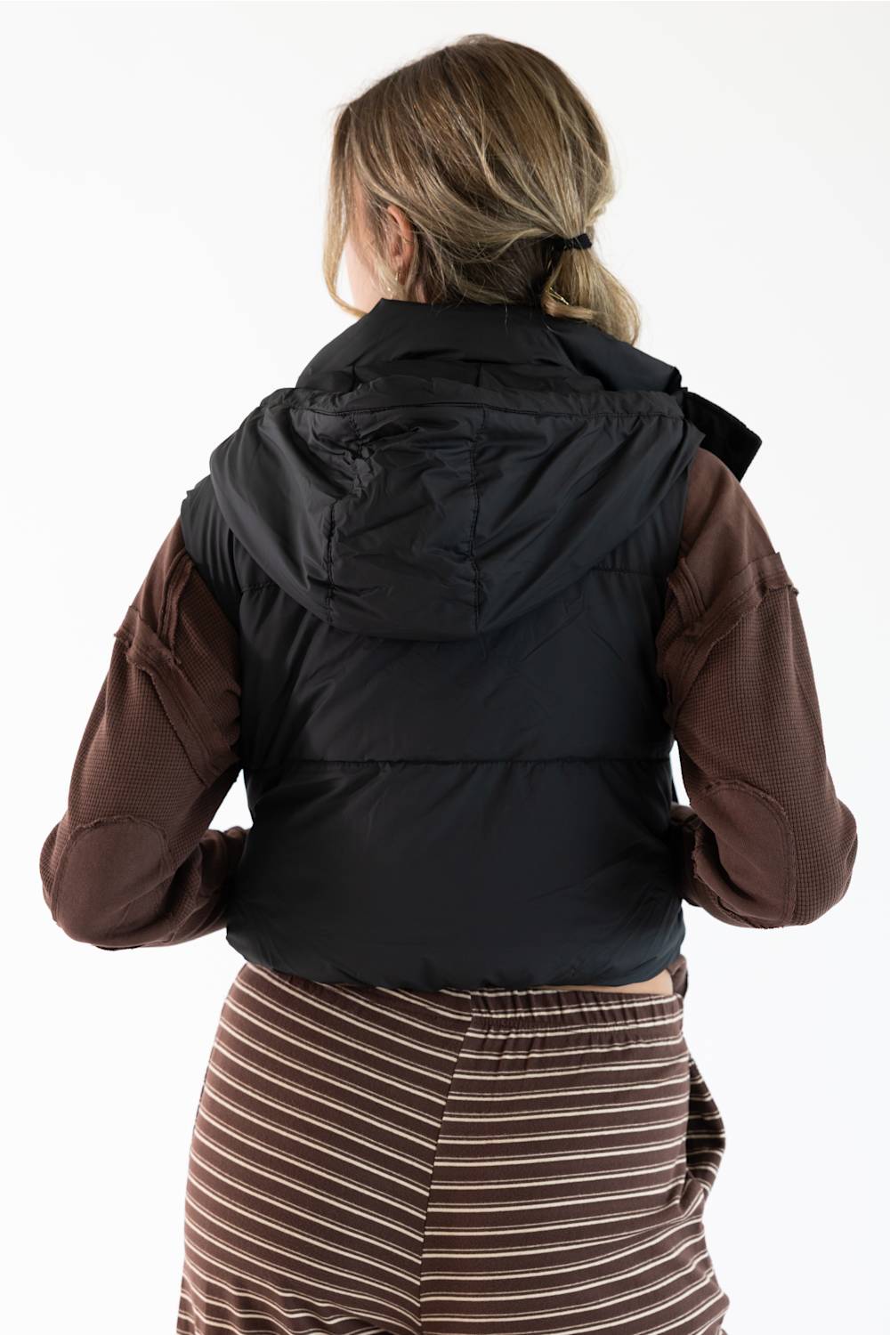 A woman viewed from behind wearing a black hooded puffer vest over brown long-sleeve sleeves and a brown-and-beige striped skirt, against a plain white studio background.