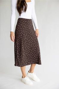 A woman wearing a brown midi skirt with a white polka dot pattern, styled with a white long-sleeve top and white sneakers against a light gray studio background.
