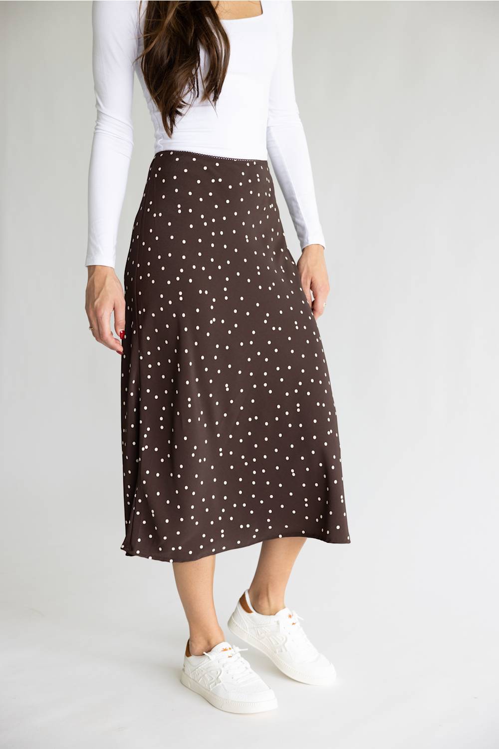 A woman wearing a brown midi skirt with a white polka dot pattern, styled with a white long-sleeve top and white sneakers against a light gray studio background.