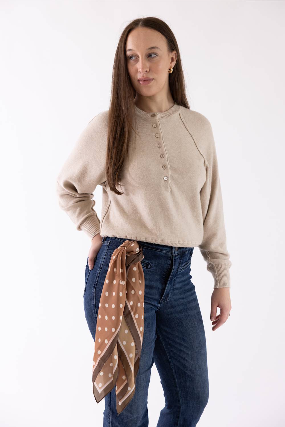 A woman modeling a beige long-sleeve henley-style knit top with brown buttons, paired with dark blue jeans and a brown polka-dot scarf tied around her waist, against a plain white studio background.