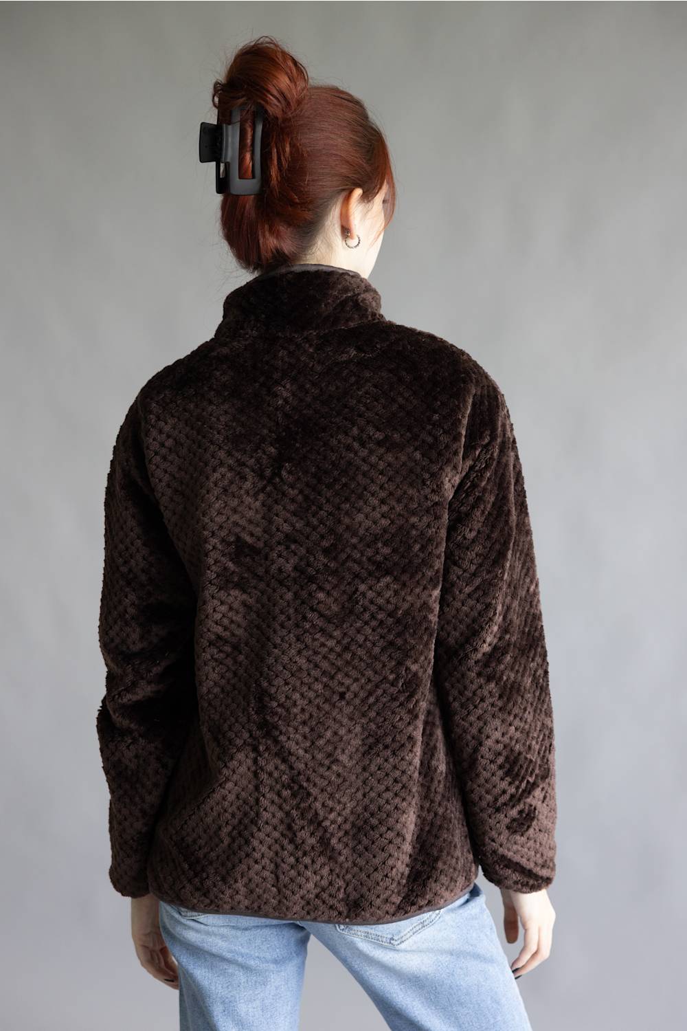 A person viewed from the back wearing a dark brown textured faux-fur jacket with a high collar and light blue jeans, set against a plain gray studio background.
