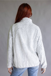 A woman with red hair is shown from the back wearing a white textured knit sweater with a high mock neck, paired with light blue jeans, against a plain gray studio background.