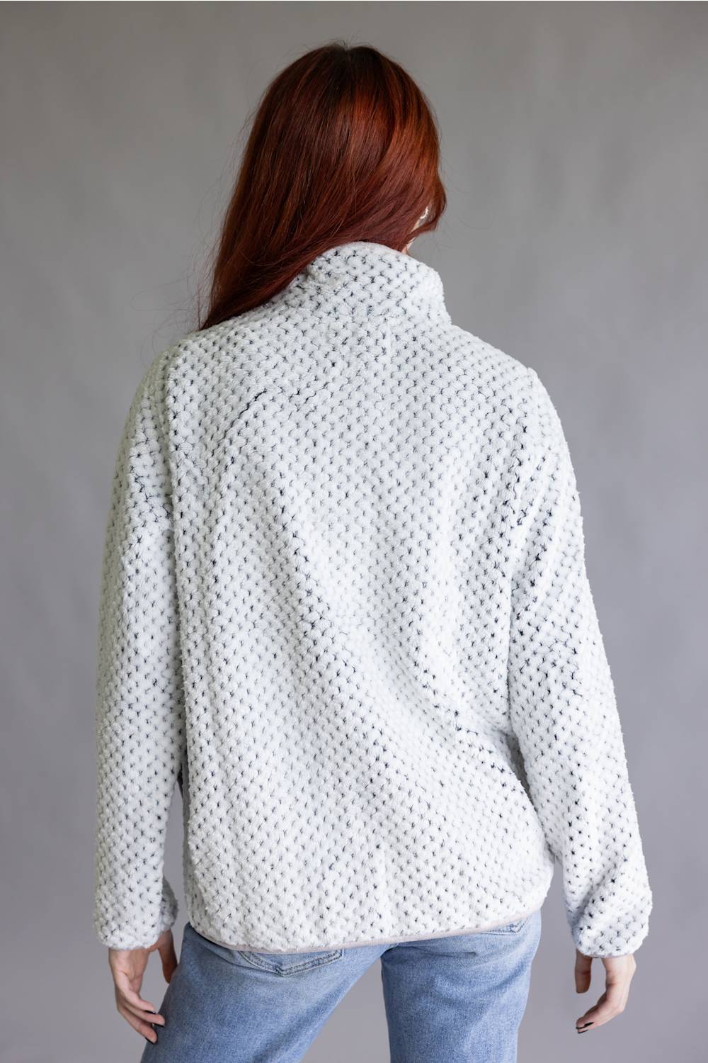 A woman with red hair is shown from the back wearing a white textured knit sweater with a high mock neck, paired with light blue jeans, against a plain gray studio background.