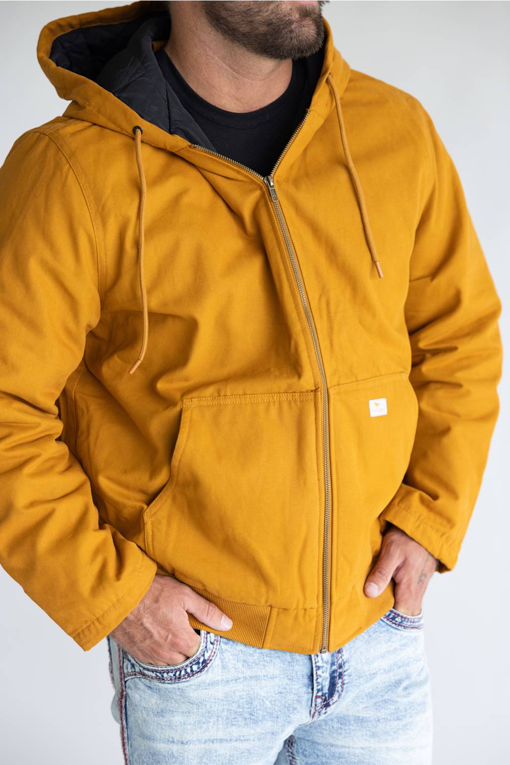 A man wearing a mustard yellow hooded jacket with a black interior lining, black zippered chest pocket, and a small white brand tag on the front, paired with light blue jeans.