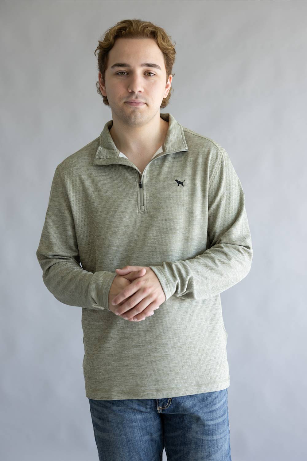 A man wearing a light sage green long-sleeve quarter-zip pullover with a small black dog logo on the left chest, paired with blue jeans, standing against a neutral gray studio background.