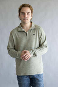 A man wearing a light sage green long-sleeve quarter-zip pullover with a small black dog logo on the left chest, paired with blue jeans, standing against a neutral gray studio background.