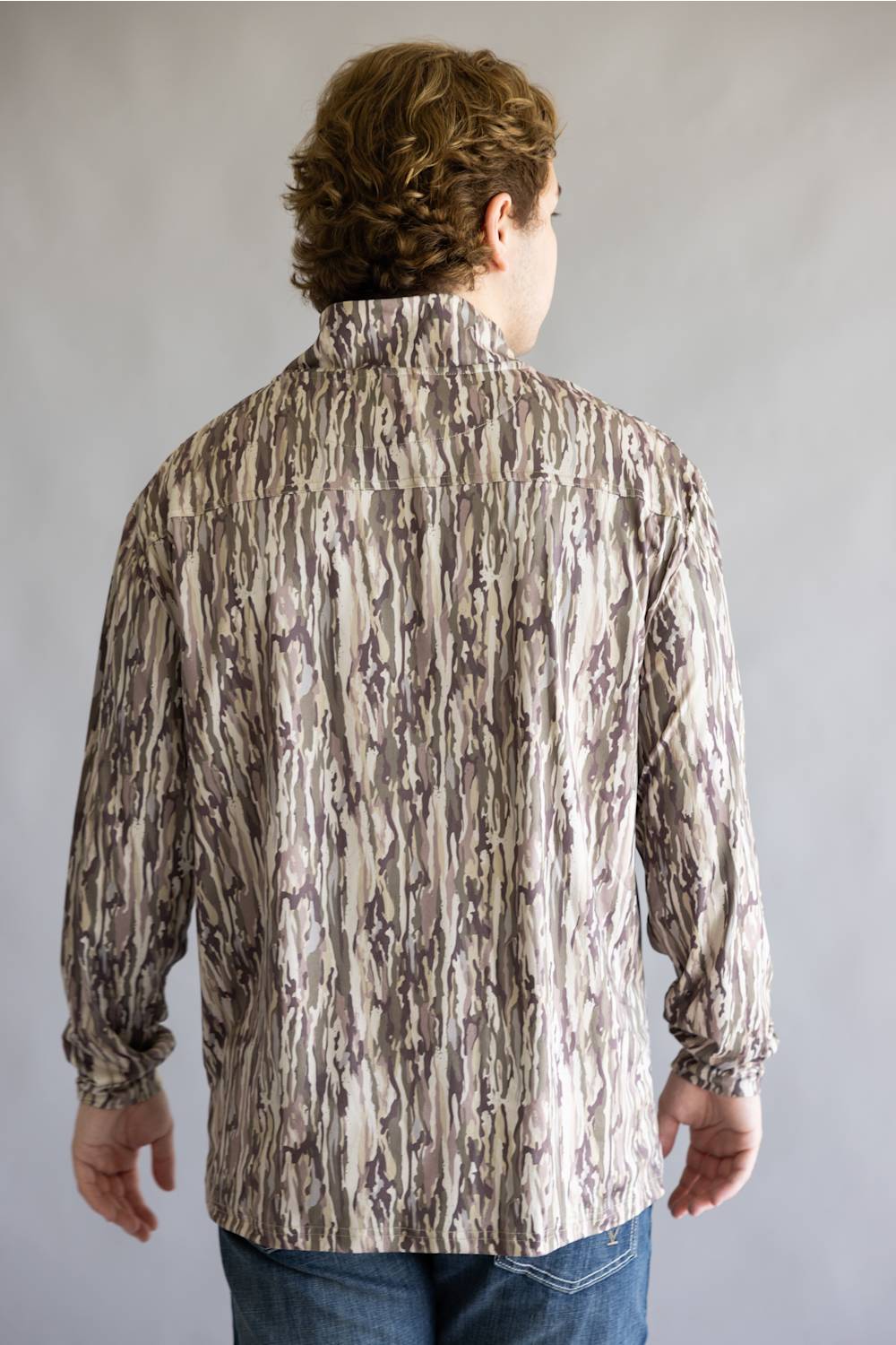 A man viewed from the back wearing a long-sleeve button-up shirt with a vertical bark-like camouflage pattern in beige, brown, and gray, paired with blue jeans against a neutral gray background.