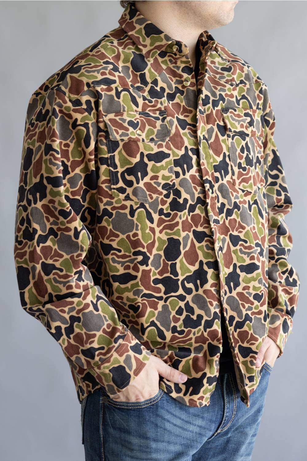 A person wearing a long-sleeve button-up shirt with an all-over camouflage pattern in brown, olive green, black, and tan, paired with blue jeans against a plain gray background.