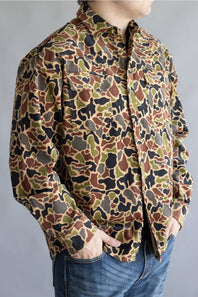 A person wearing a long-sleeve button-up shirt with an all-over camouflage pattern in brown, olive green, black, and tan, paired with blue jeans against a plain gray background.