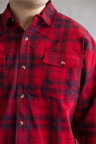 A close-up of a red and black plaid button-down shirt worn by a model, showing the collar, chest pocket with a snap button, and fabric texture.